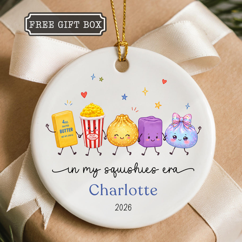 May include: A round white ceramic ornament featuring a gold hanging string and a graphic of five cartoon characters including butter, popcorn, a dumpling, a purple block, and a blue pouch. Text reads "in my squishies era", "Charlotte", and "2026".