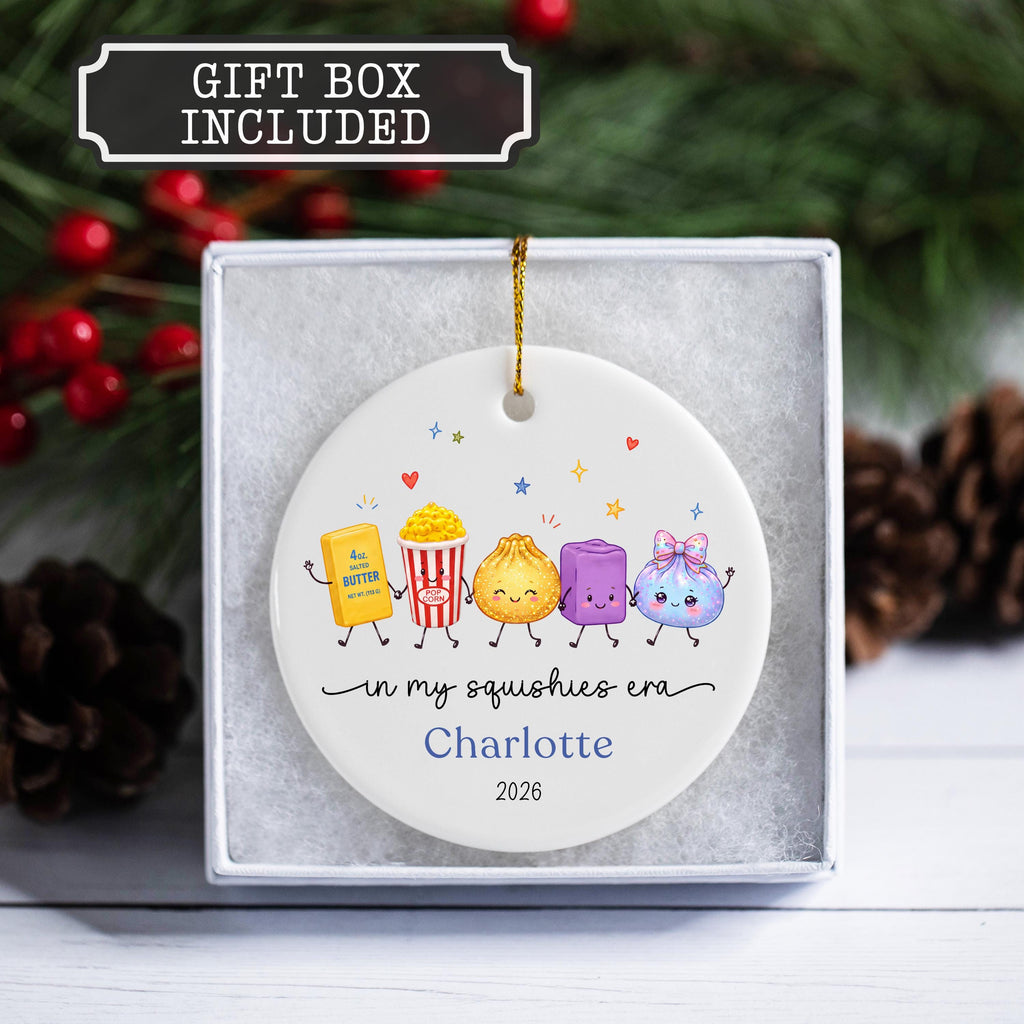 May include: A white circular ceramic ornament featuring a gold hanging string, decorated with colorful cartoon characters of butter, popcorn, a dumpling, a purple cube, and a blue pouch. Text reads: in my squishies era, Charlotte, 2026.