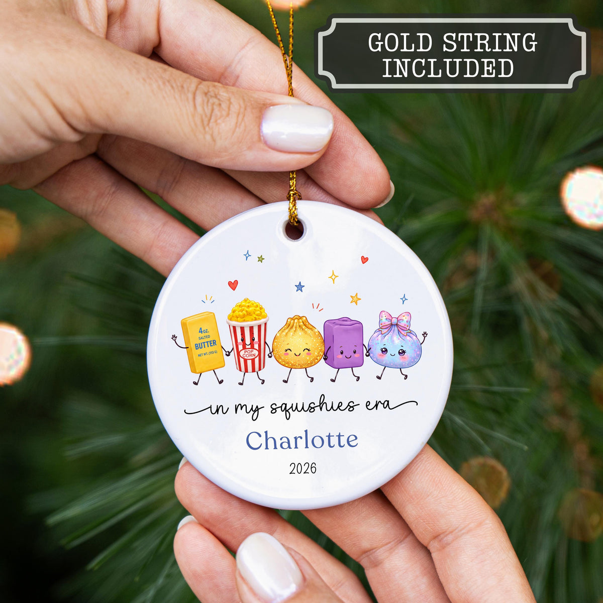 May include: A round white ceramic ornament featuring a colorful illustration of five cartoon characters including a butter stick, popcorn, dumpling, purple square, and a blue pouch. The text reads in my squishies era, Charlotte, and 2026.