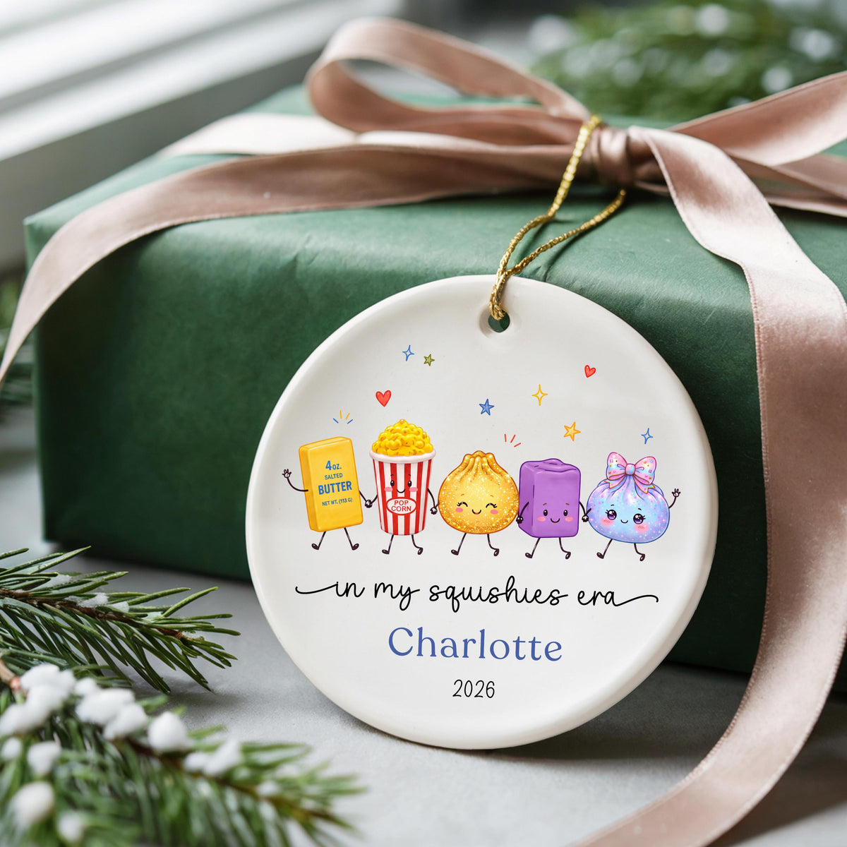 May include: A round white ceramic ornament featuring a colorful illustration of five cute cartoon characters including a butter stick, popcorn, dumpling, purple block, and a bow-tied pouch. Text reads: in my squishies era, Charlotte, 2026.
