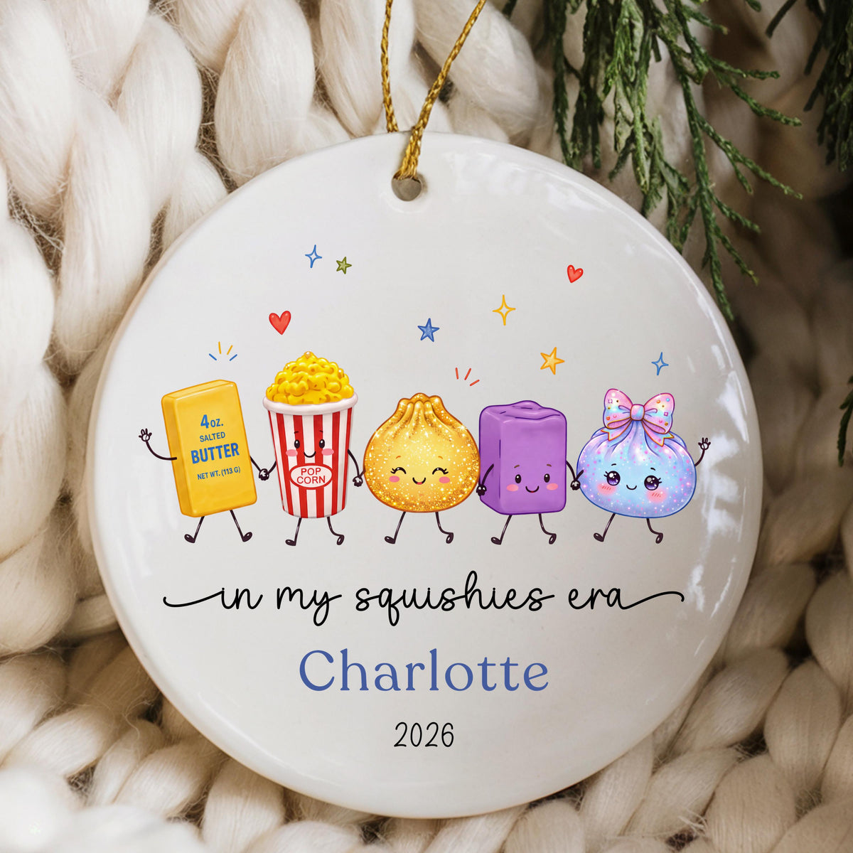May include: A round white ceramic ornament featuring a gold hanging string. The design shows five cartoon characters holding hands, including butter, popcorn, a dumpling, a purple block, and a blue pouch. Text reads: in my squishies era, Charlotte, 2026.