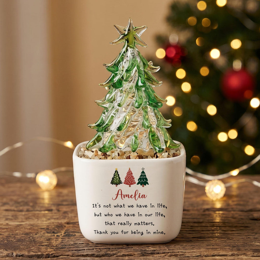 May include: A clear and green glass Christmas tree ornament with a star topper, set in a white square ceramic planter filled with small stones. The planter features three small tree icons and the text: Amelia, It's not what we have in life, but who we have in our life, that really matters, Thank you for being in mine.