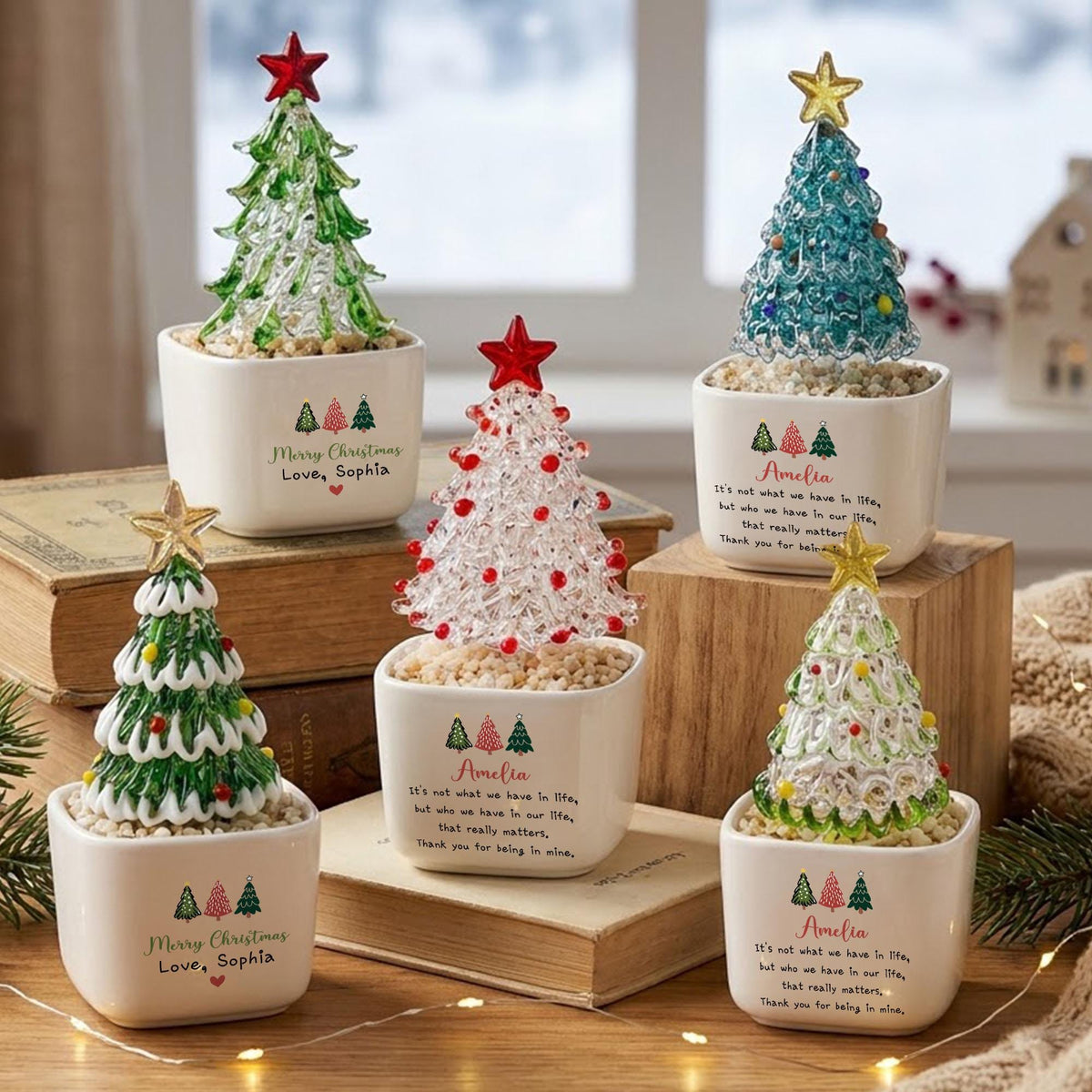 May include: Five glass Christmas tree figurines in white ceramic pots. Each tree features a star topper and sits on a bed of small stones. The pots are printed with text including Merry Christmas, Love, Sophia, and Amelia, along with tree icons.
