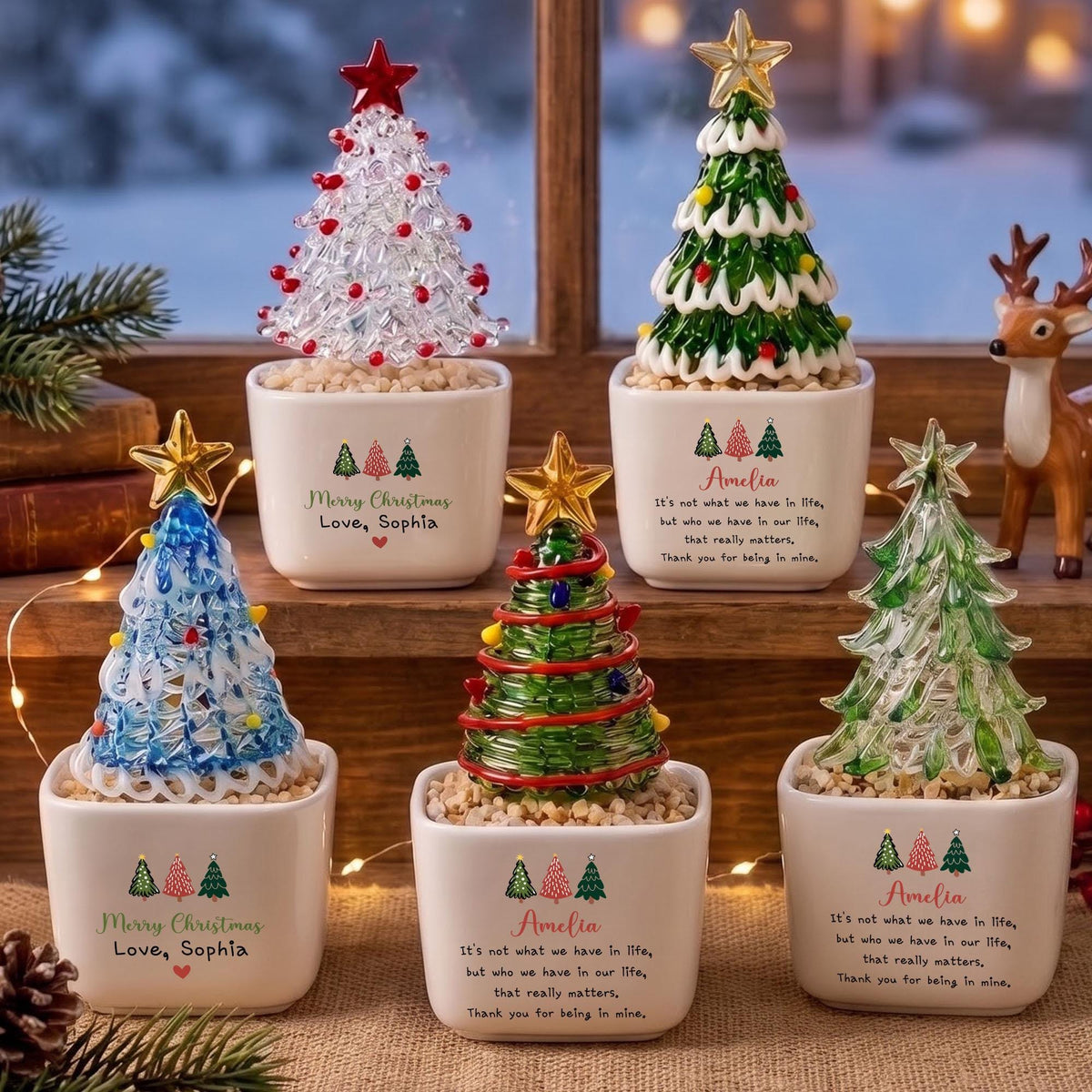 May include: Five decorative glass Christmas trees in white ceramic pots. The trees feature various colors including clear, green, and blue with star toppers. The pots are printed with text such as Merry Christmas, Love, Sophia, and Amelia.