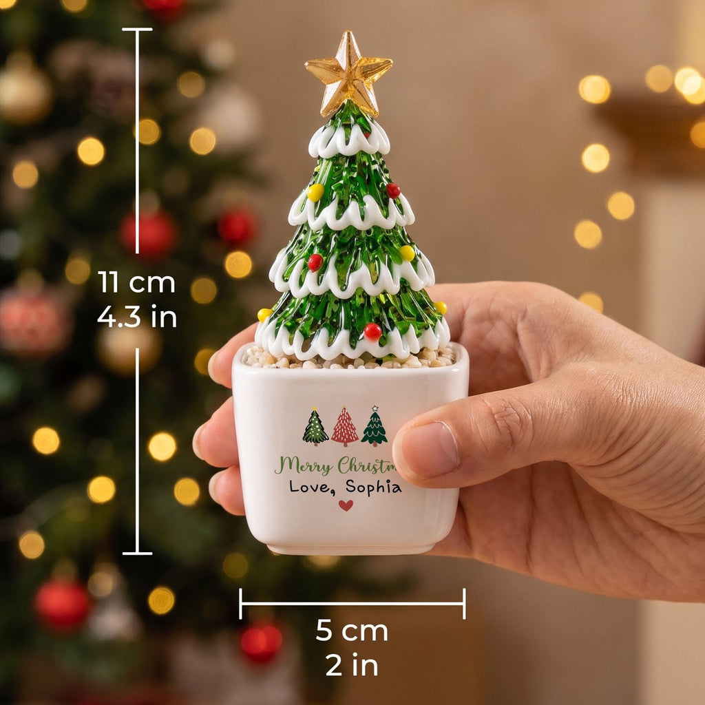May include: A green glass Christmas tree figurine with white trim and colorful accents, topped with a gold star. It sits in a white square pot labeled Merry Christmas Love, Sophia. The item measures 4.3 inches tall and 2 inches wide.