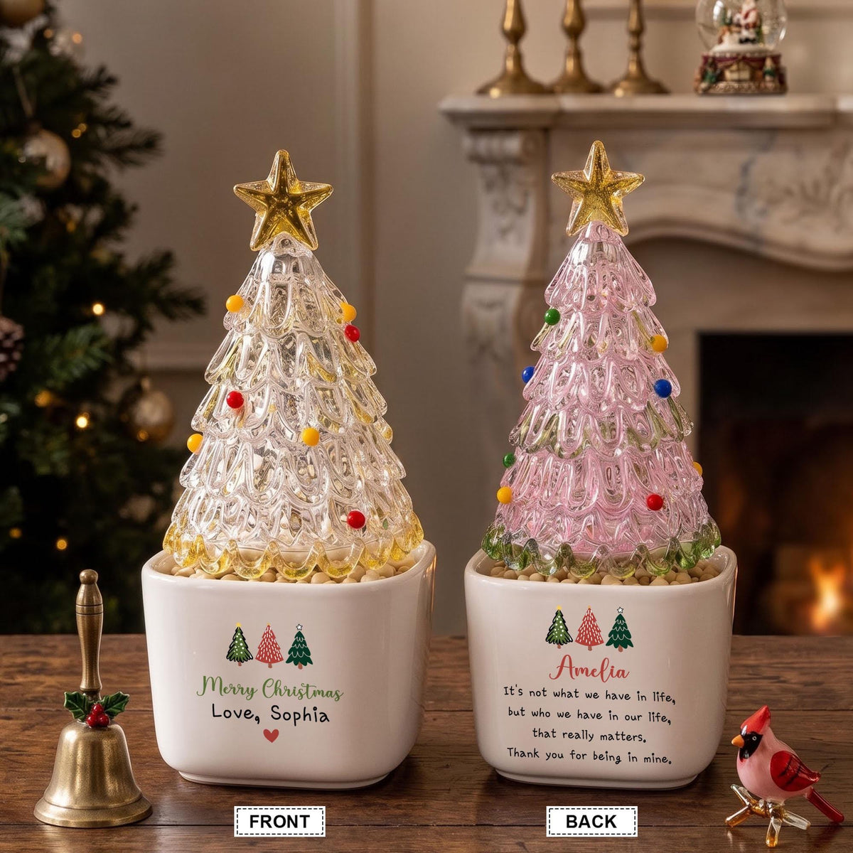 May include: Two glass Christmas tree figurines in white ceramic pots. The left tree is clear with yellow, red, and white ornaments, labeled Merry Christmas Love, Sophia. The right tree is pink with colorful ornaments, labeled Amelia and a quote.