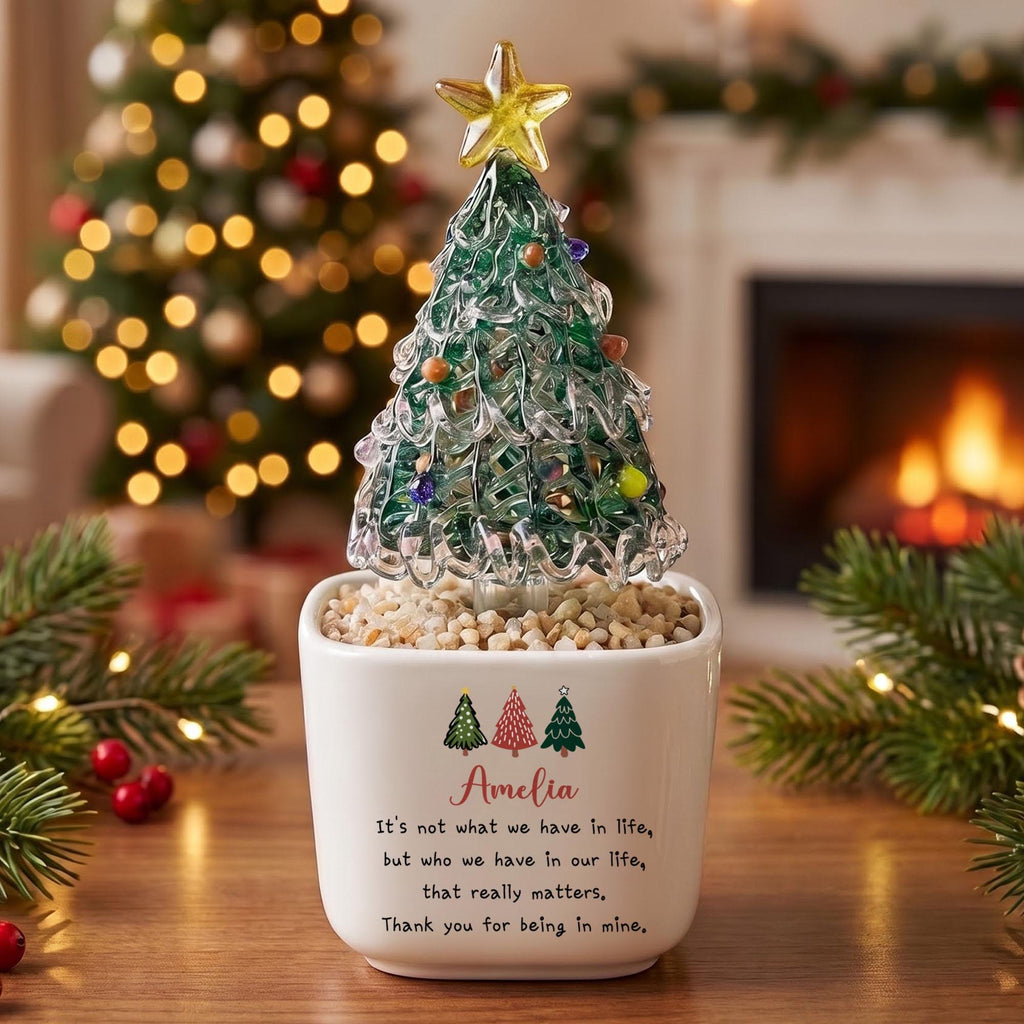 May include: A glass Christmas tree figurine with a yellow star topper, set in a white ceramic planter filled with small stones. The planter features three small tree icons and the text: Amelia, It's not what we have in life, but who we have in our life, that really matters, Thank you for being in mine.