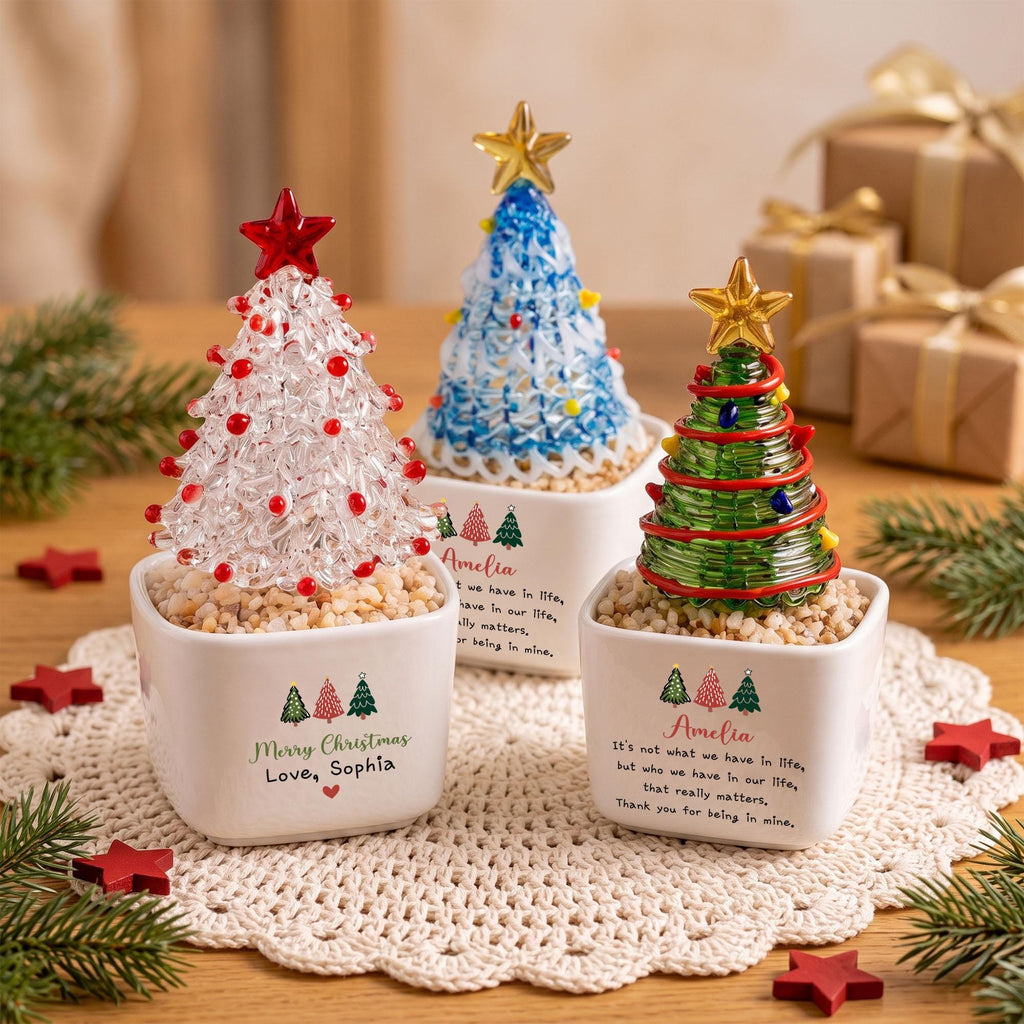 May include: Three decorative glass Christmas trees in white square ceramic pots. One tree is clear with red accents, one is blue with multi-colored accents, and one is green with red and yellow accents. Each pot features printed text and tree icons.