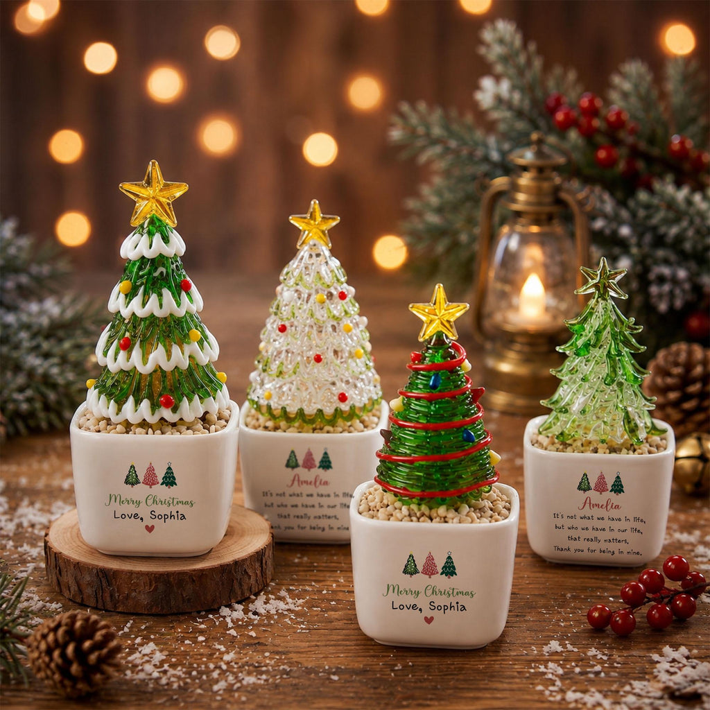 May include: Four miniature glass Christmas trees in white square ceramic pots. Each tree features a yellow star topper and colorful ornaments. The pots are printed with the text Merry Christmas, Love, Sophia or Amelia and small tree icons.