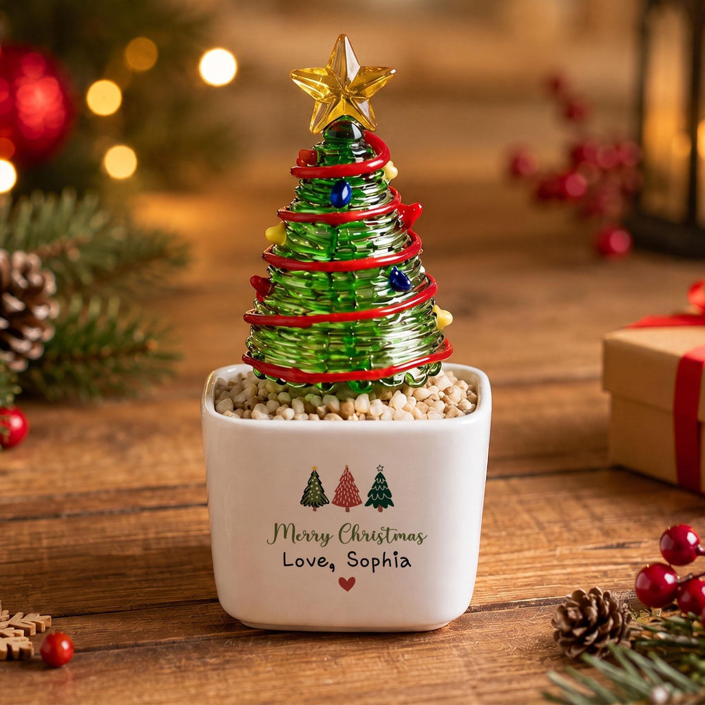 May include: A glass Christmas tree figurine with a yellow star topper, red garland, and colorful accents, set in a white square ceramic planter filled with small stones. The planter features three small tree icons and the text Merry Christmas Love, Sophia.