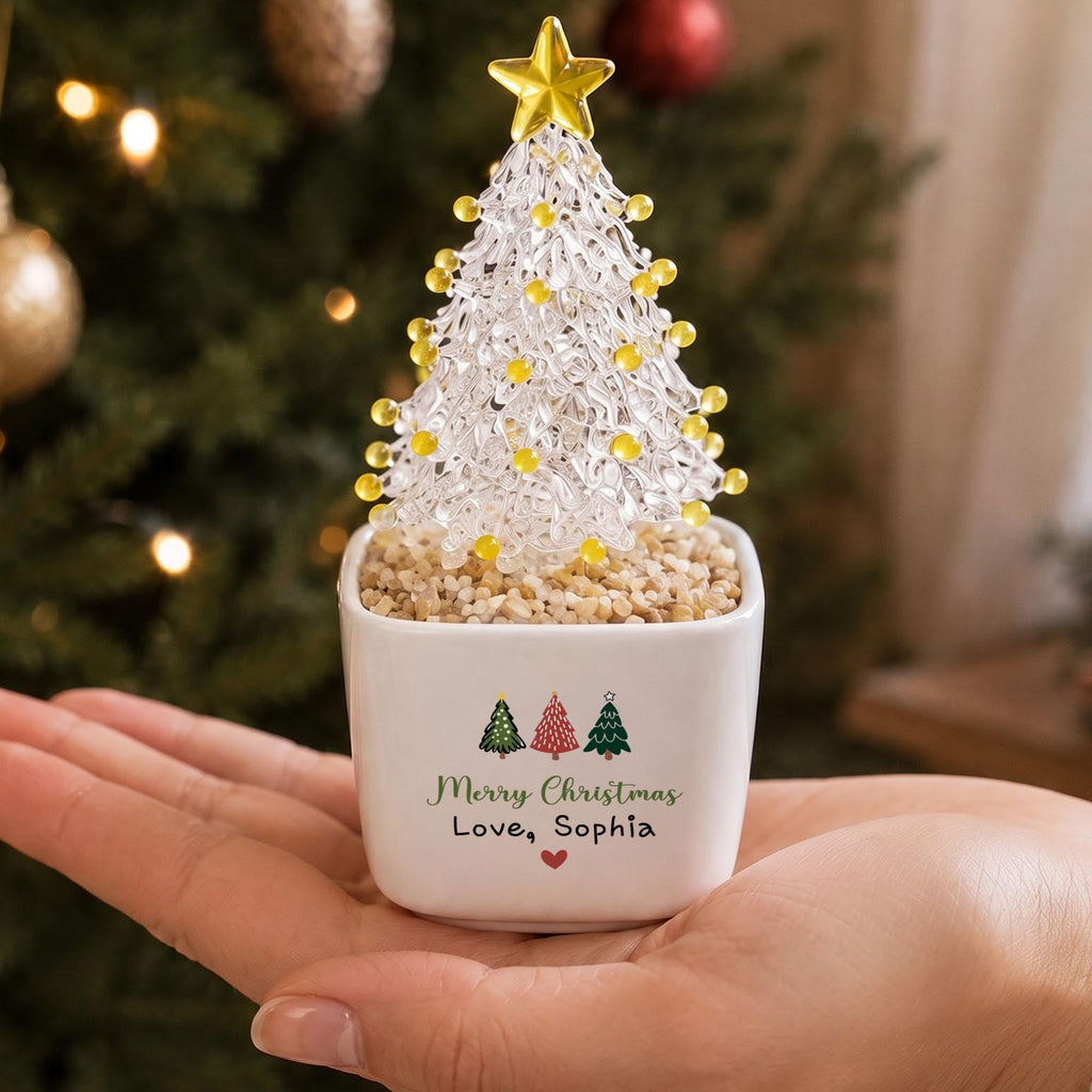 May include: A clear glass Christmas tree figurine with yellow glass bead ornaments and a yellow star topper, set in a white square ceramic pot filled with small tan pebbles. The pot features three small tree icons and the text Merry Christmas Love, Sophia.