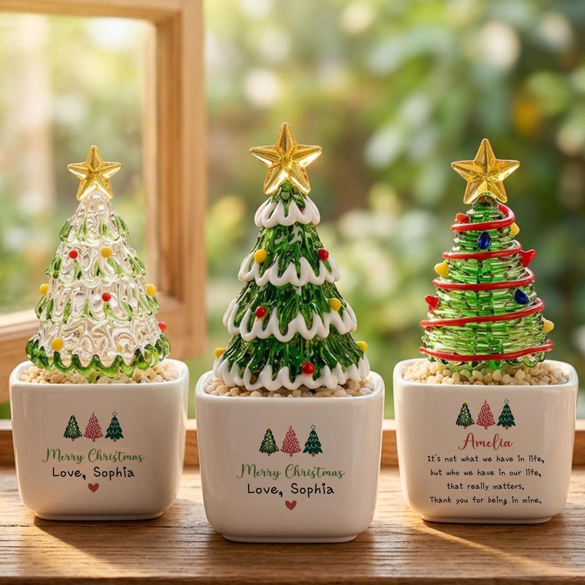 May include: Three glass Christmas tree figurines in white square ceramic pots. The trees feature gold stars on top and colorful accents. The pots are printed with the text Merry Christmas, Love, Sophia, and a quote about Amelia, along with small tree icons.