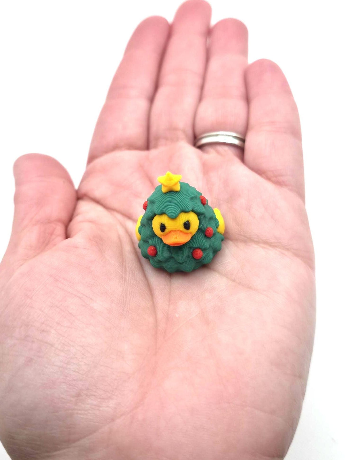 May include: A small rubber duck figurine shaped like a Christmas tree, featuring a green textured body, a yellow star on top, small red circular ornaments, and a yellow beak with black eyes, designed as a festive holiday decoration.