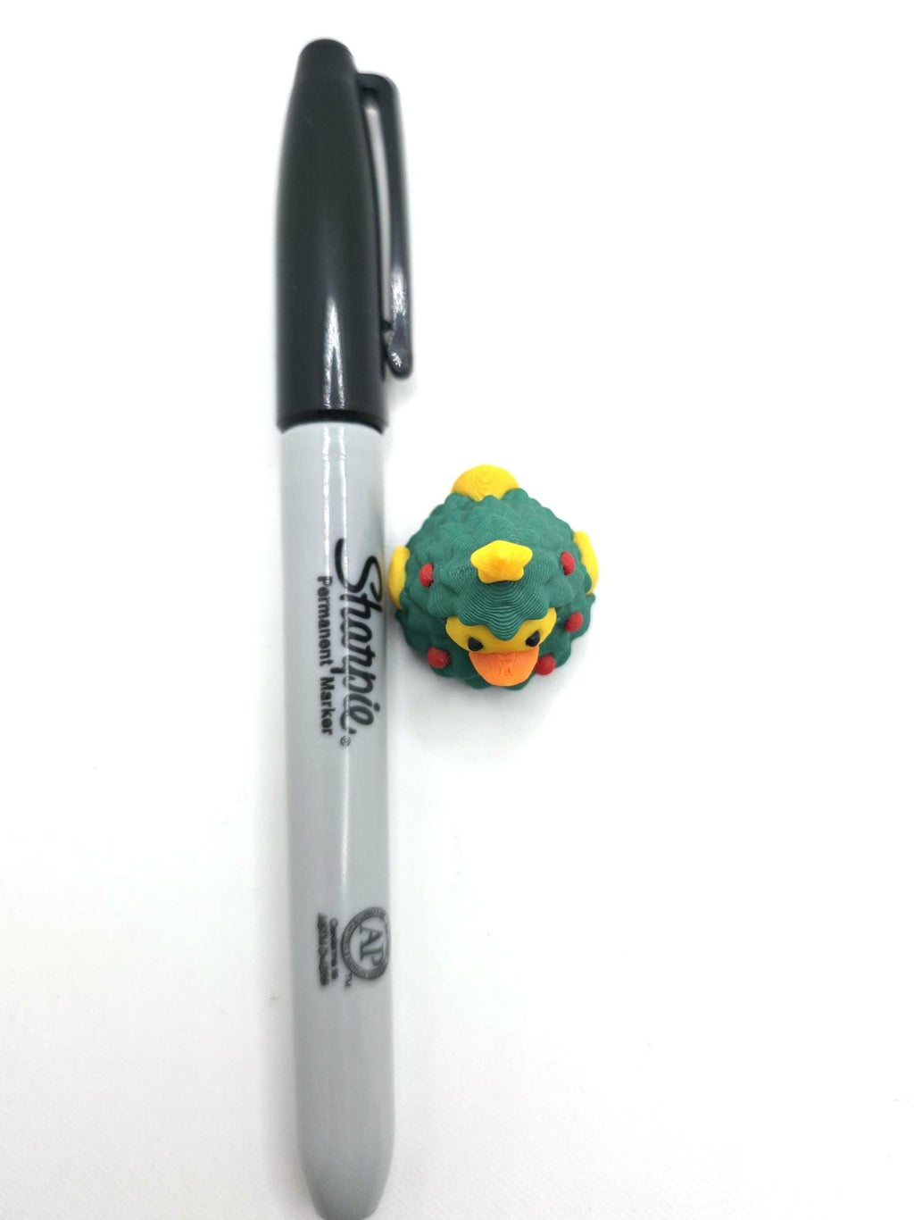 May include: A small rubber duck shaped like a Christmas tree with green foliage, red ornaments, and a yellow star on top, positioned next to a standard black Sharpie permanent marker for scale to show its compact size.