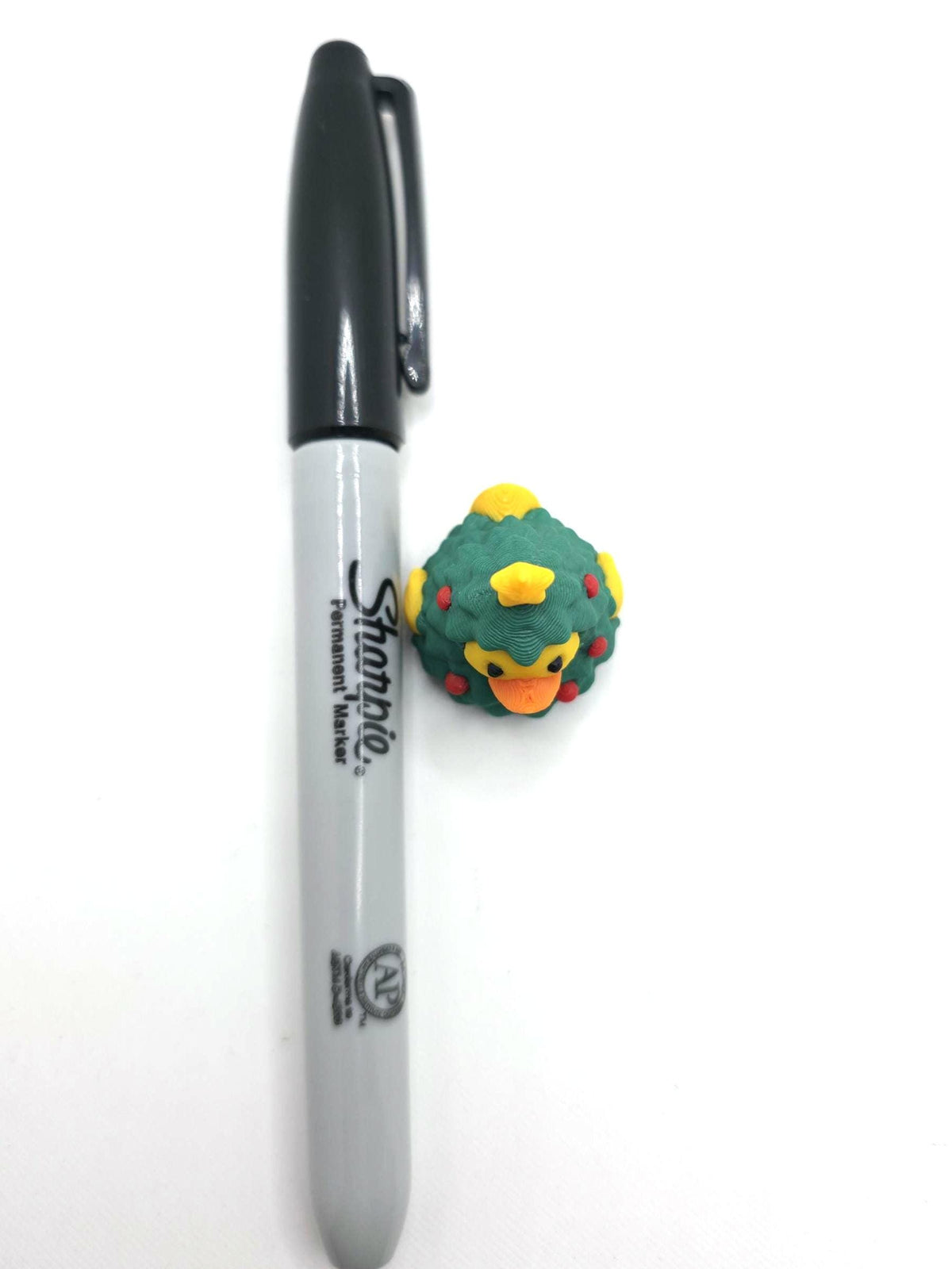 May include: A small rubber duck shaped like a Christmas tree with green foliage, red ornaments, and a yellow star on top, positioned next to a standard black Sharpie permanent marker for scale to show its compact size.