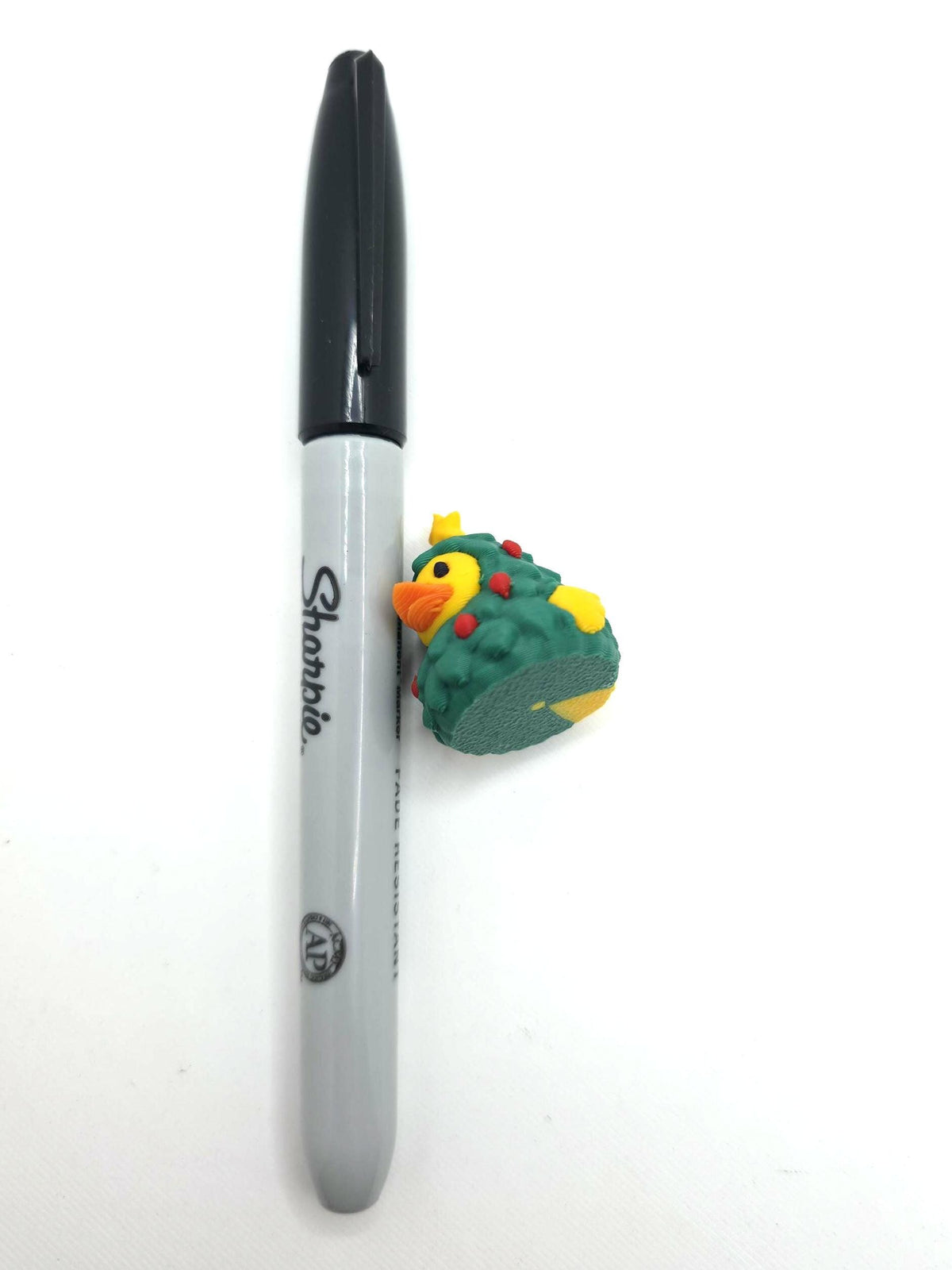 May include: A gray Sharpie permanent marker with a black cap positioned next to a small rubber duck figurine wearing a green Christmas tree costume with red ornaments and a yellow star on top, showing the scale of the duck against the marker.