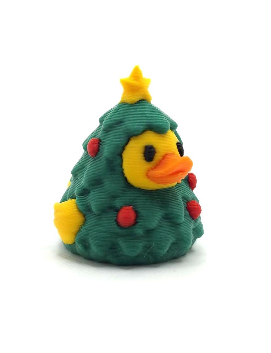 May include: A 3D printed figurine shaped like a yellow duck wearing a dark green Christmas tree costume. The duck has an orange beak and black eyes, while the tree features a yellow star on top and small red circular ornaments throughout.