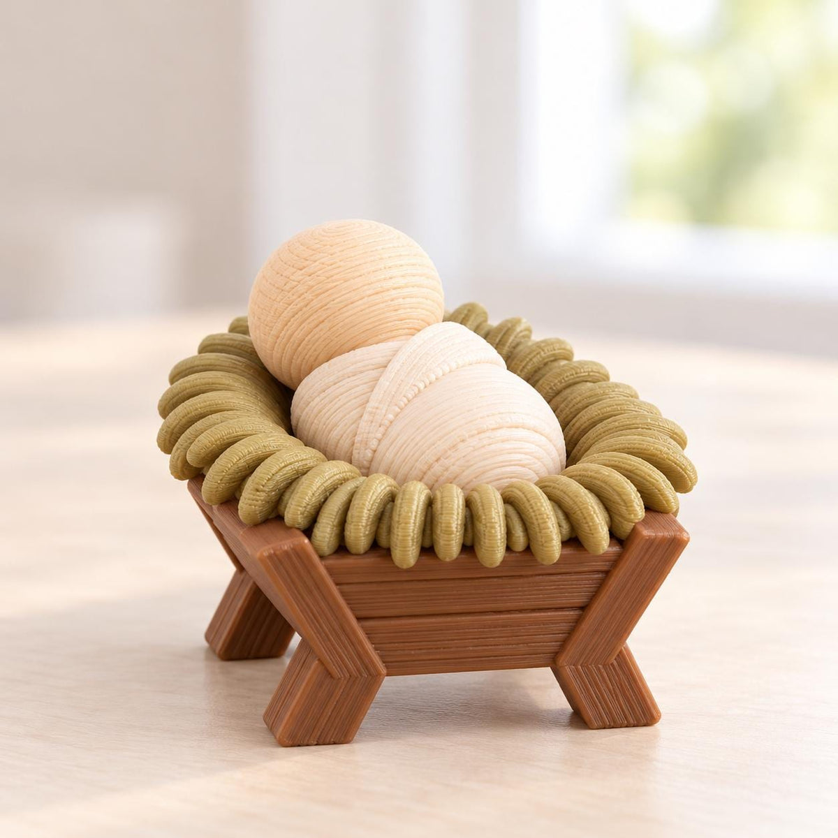 May include: A small wooden manger figurine featuring a light tan rounded head and a swaddled body resting inside a brown cradle. The cradle is surrounded by a textured olive green ring that mimics hay, all crafted with visible wood grain details.