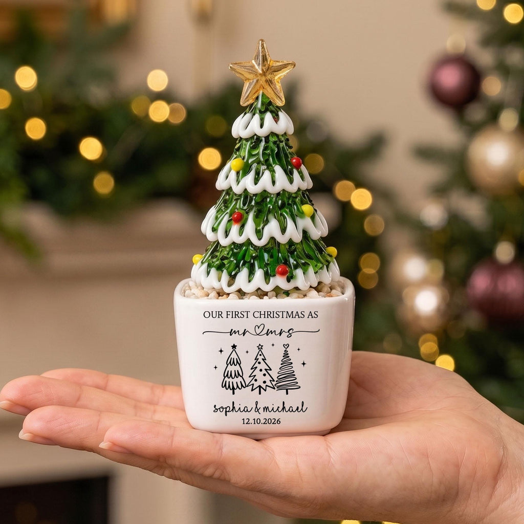 May include: A glass Christmas tree figurine with green branches, white snow-like trim, and small colorful ornaments, topped with a gold star. The white ceramic base features the text "OUR FIRST CHRISTMAS AS mr & mrs", "sophia & michael", and "12.10.2026".