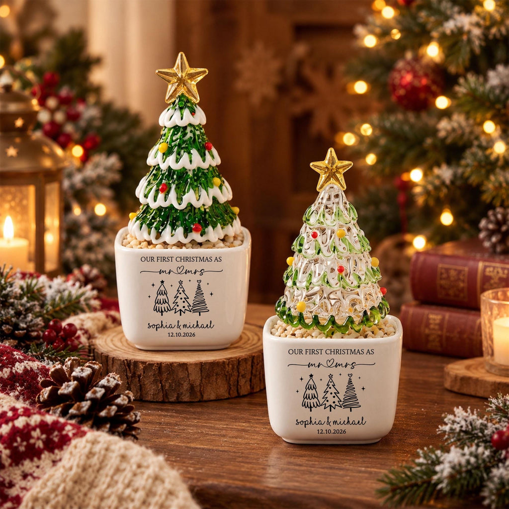 May include: Two white ceramic square planters containing miniature Christmas trees. The left tree is green with white snow-like trim and red accents. The right tree is clear glass with green and yellow accents. Both feature gold stars and text: OUR FIRST CHRISTMAS AS MR & MRS, SOPHIA & MICHAEL, 12.10.2026.