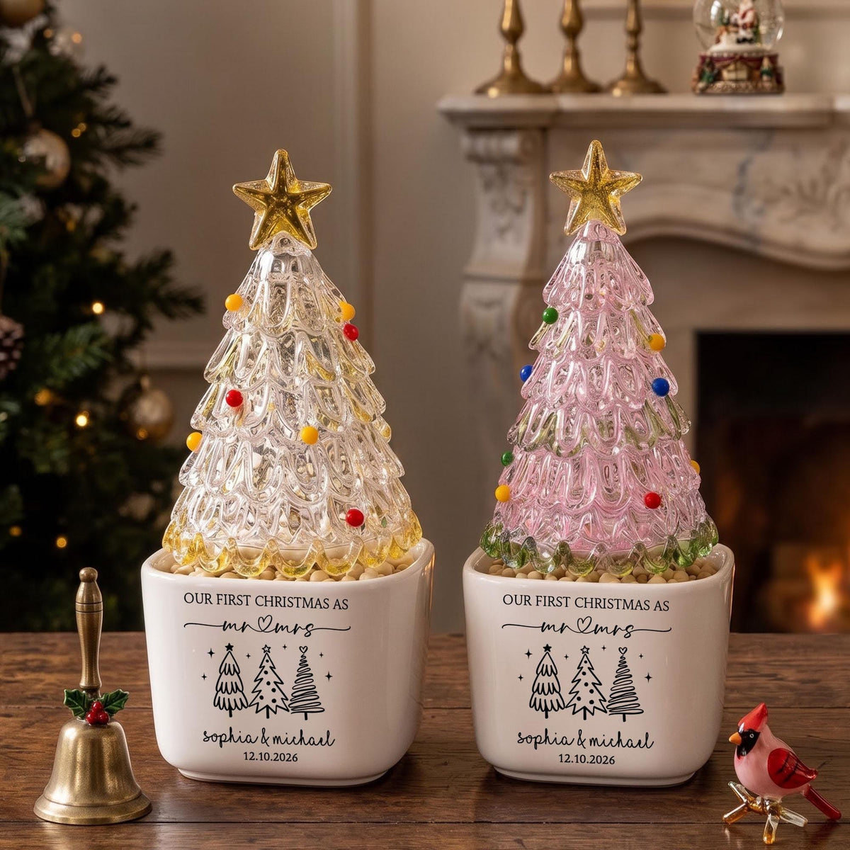 May include: Two decorative glass Christmas trees in white ceramic planters. One tree is clear glass, the other is pink. Both feature a gold star topper and colorful glass ornaments. The planters are printed with "OUR FIRST CHRISTMAS AS MR & MRS", "SOPHIA & MICHAEL", and "12.10.2026".
