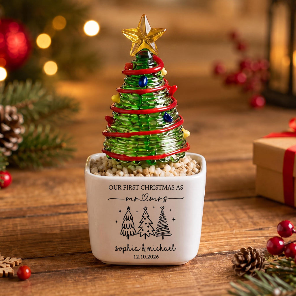 May include: A glass Christmas tree figurine with a yellow star topper, red garland, and colorful ornaments, set in a white square ceramic planter filled with small stones. The planter features black text reading Our First Christmas as Mr & Mrs, Sophia & Michael, 12.10.2026, and three tree illustrations.