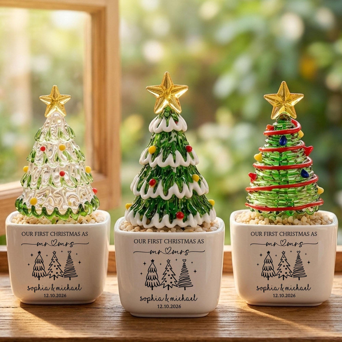 May include: Three decorative glass Christmas tree ornaments in white square ceramic pots. Each pot features the text "OUR FIRST CHRISTMAS AS mr & mrs", "SOPHIA & MICHAEL", and "12.10.2026" with black tree illustrations. The trees are green or clear glass with gold stars.