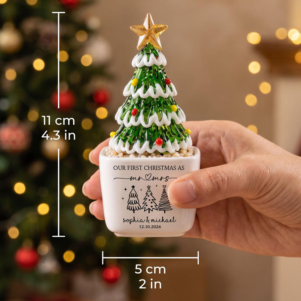 May include: A glass Christmas tree ornament with a gold star topper, green branches, white trim, and colorful accents, set in a white square base. The base features the text "OUR FIRST CHRISTMAS AS mr & mrs", "SOPHIA & MICHAEL", "12.10.2026", and tree icons. Height is 4.3 inches, width is 2 inches.