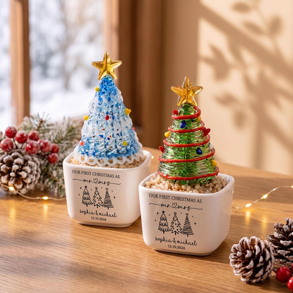 May include: Two white square ceramic planters containing glass Christmas trees. One tree is blue and white, the other is green with red accents. Both are topped with gold stars and feature the text "OUR FIRST CHRISTMAS AS mr & mrs" with names.