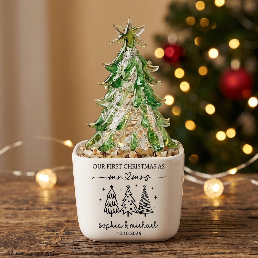 May include: A clear and green glass Christmas tree ornament with a star topper, set in a white square ceramic planter. The planter features the text "OUR FIRST CHRISTMAS AS mr & mrs", "sophia & michael", "12.10.2026", and three tree icons.