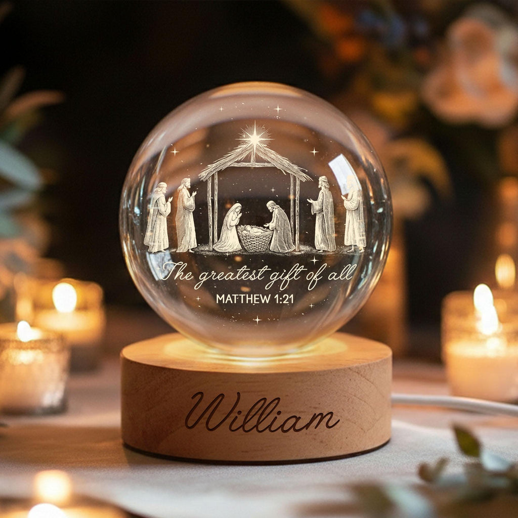 May include: A clear glass sphere featuring an etched nativity scene with a manger, figures, and a star, inscribed with "The greatest gift of all" and "MATTHEW 1:21". The sphere rests on a cylindrical wooden base engraved with the name "William".