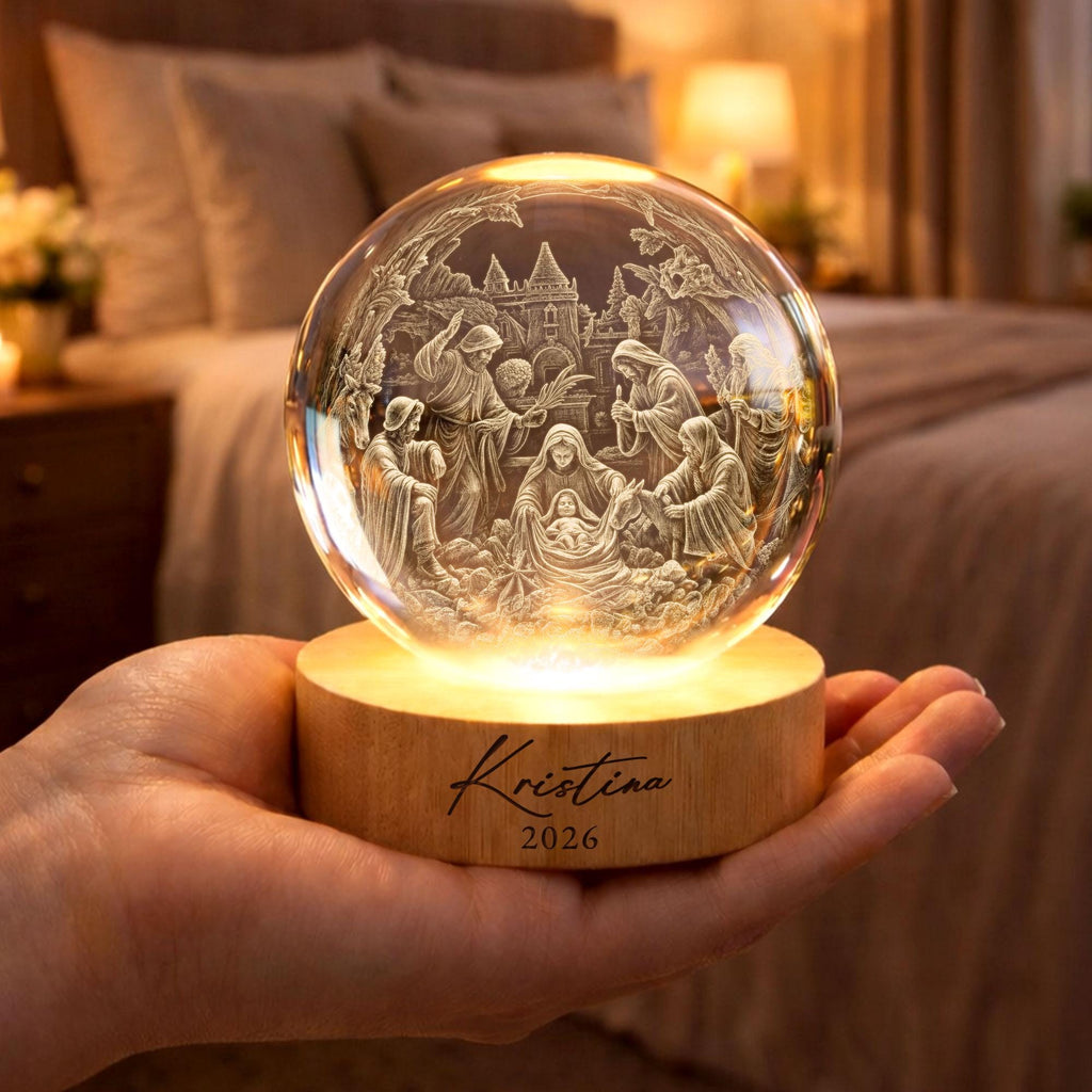 May include: A clear glass sphere featuring an intricate 3D laser-engraved nativity scene with figures and a castle. The sphere rests on a round, light-colored wooden base illuminated from below, with the name Kristina and the year 2026 engraved.