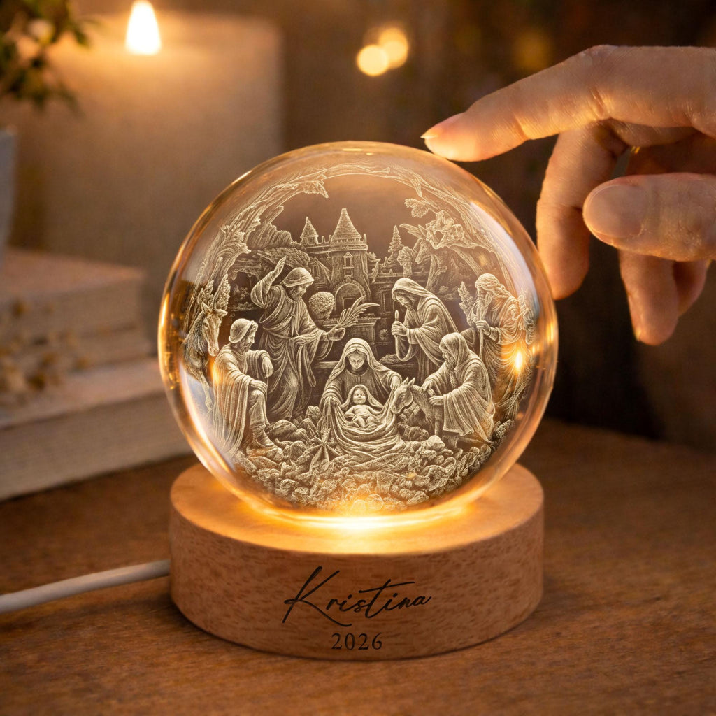 May include: A clear glass sphere featuring an intricate 3D laser-engraved nativity scene with figures and a building. The sphere rests on a round wooden base with the engraved text "Kristina" and "2026" centered on the front surface.