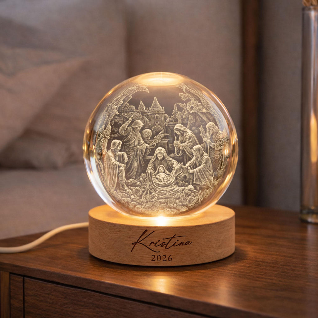 May include: A clear glass sphere lamp featuring an intricate 3D laser-engraved nativity scene inside. The sphere rests on a round, light-colored wooden base with the name Kristina and the year 2026 engraved in black script on the front surface.