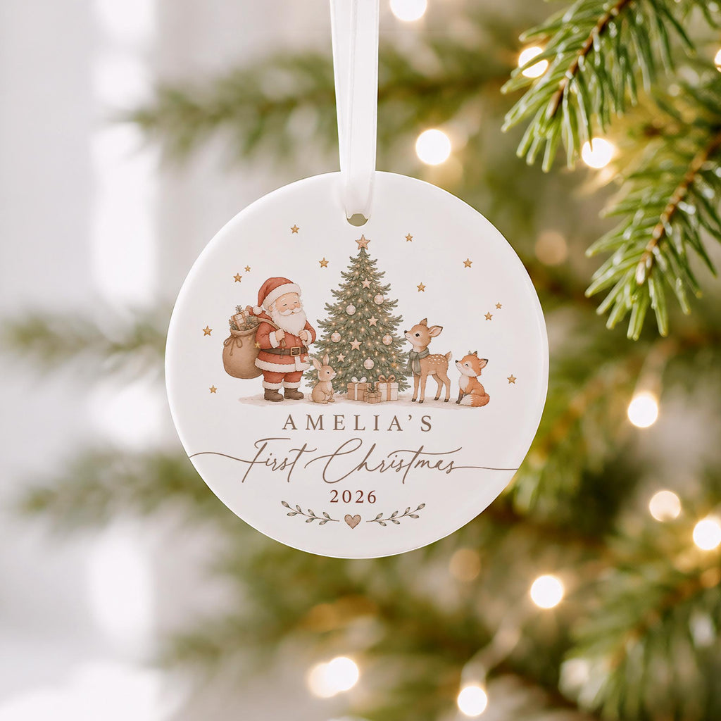 May include: A round white ceramic ornament hanging from a white ribbon. The design features a Christmas tree, Santa Claus, a rabbit, a deer, and a fox. Text reads AMELIA'S First Christmas 2026 with small stars and a heart accent below the date.