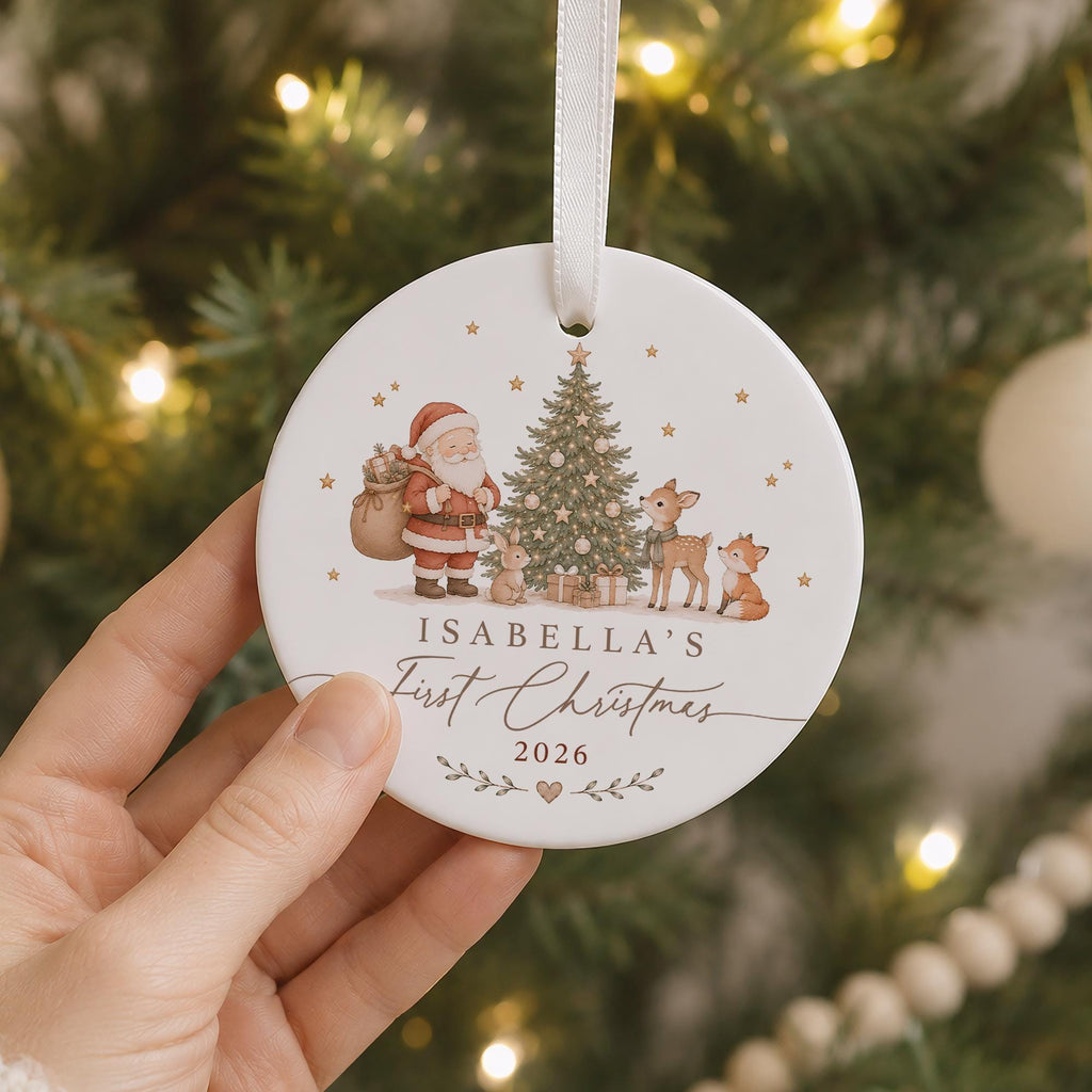 May include: A round white ceramic Christmas ornament with a white ribbon hanger. The design features Santa Claus, a decorated tree, a rabbit, a deer, and a fox. Text reads: ISABELLA'S First Christmas 2026, accented with small gold stars and leaves.