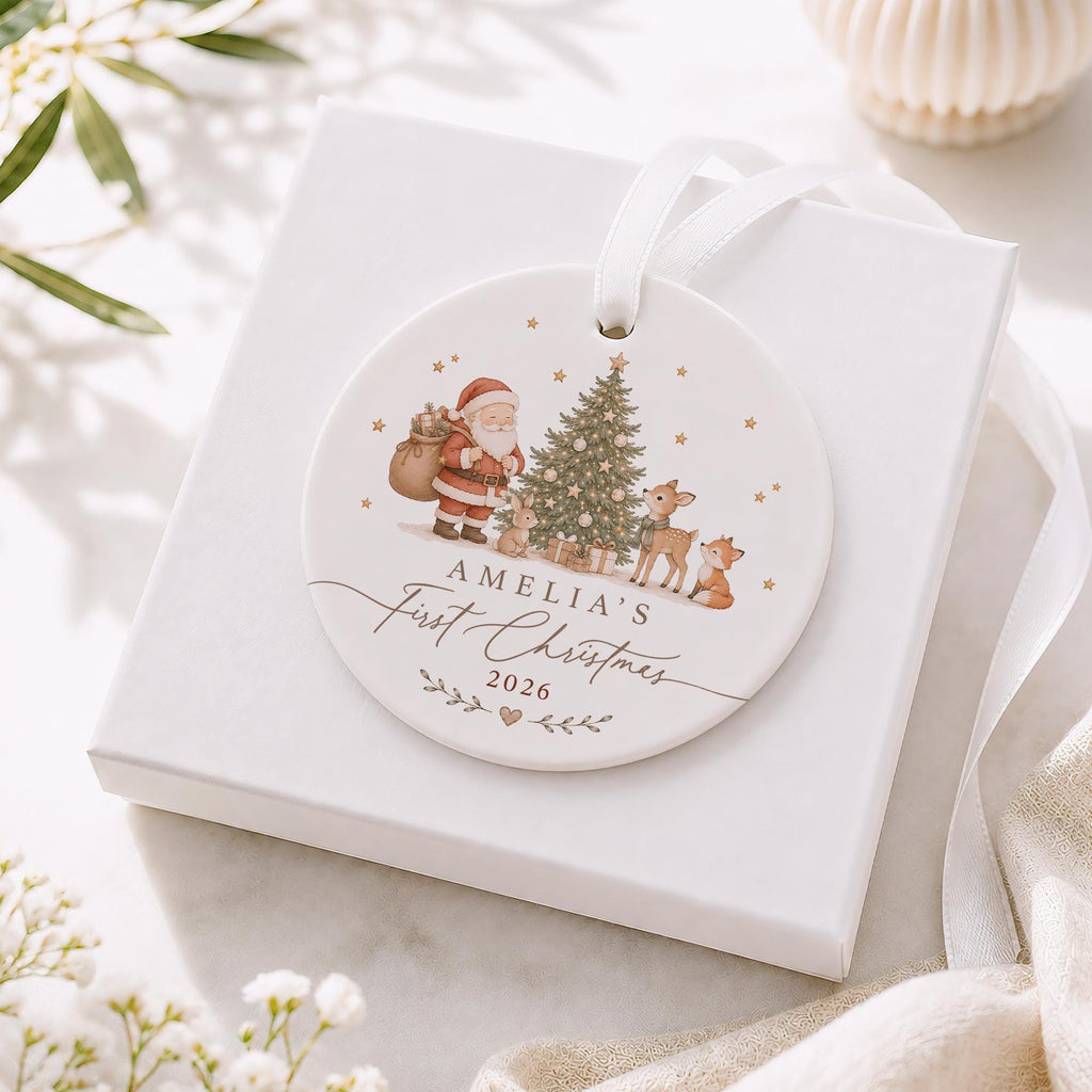 May include: A round white ceramic Christmas ornament featuring a printed illustration of Santa Claus, a decorated tree, a rabbit, a deer, and a fox. The text reads Amelia's First Christmas 2026. It includes a white ribbon for hanging.