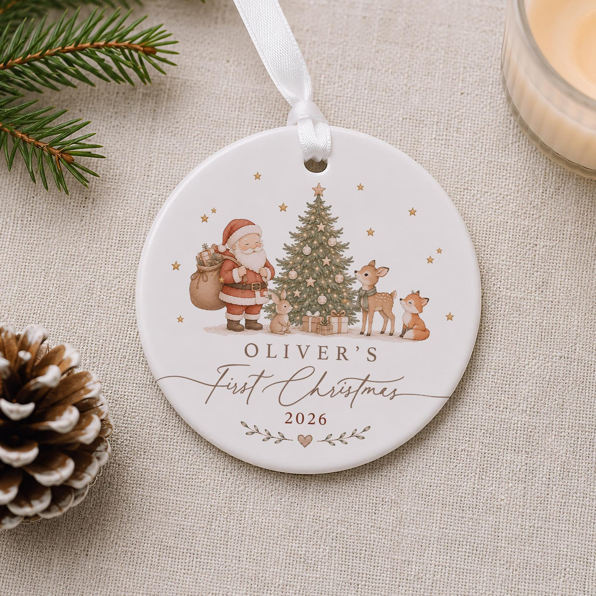 May include: A round white ceramic Christmas ornament hanging from a white ribbon. The ornament features a printed illustration of Santa Claus, a decorated Christmas tree, a rabbit, a deer, and a fox. Text reads OLIVER'S First Christmas 2026.