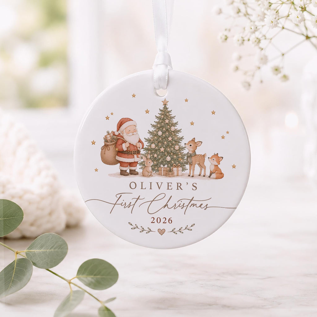 May include: A round white ceramic ornament hanging from a white ribbon. The front features a printed illustration of Santa Claus, a decorated Christmas tree, a rabbit, a deer, and a fox. Text reads: OLIVER'S First Christmas 2026.