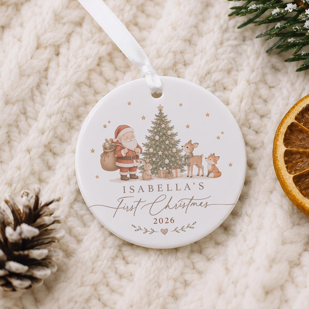 May include: A round white ceramic ornament with a white ribbon, featuring a printed illustration of Santa Claus, a Christmas tree, a deer, a rabbit, and a fox. The text reads: ISABELLA'S First Christmas 2026, decorated with small stars and a heart.