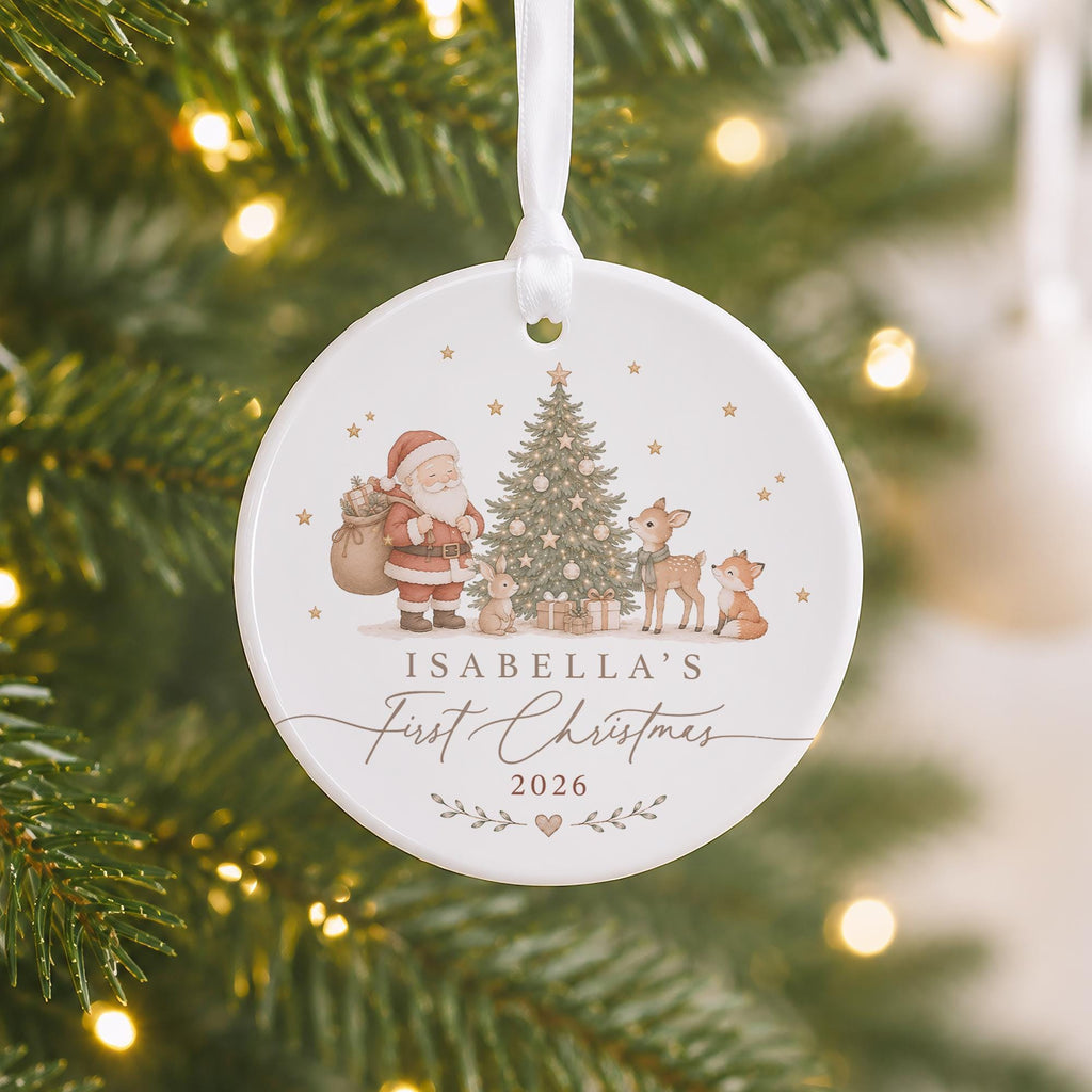 May include: A round white ceramic Christmas ornament hanging from a white ribbon. The front features a printed illustration of Santa Claus, a decorated tree, a rabbit, a deer, and a fox. Text reads: ISABELLA'S First Christmas 2026.