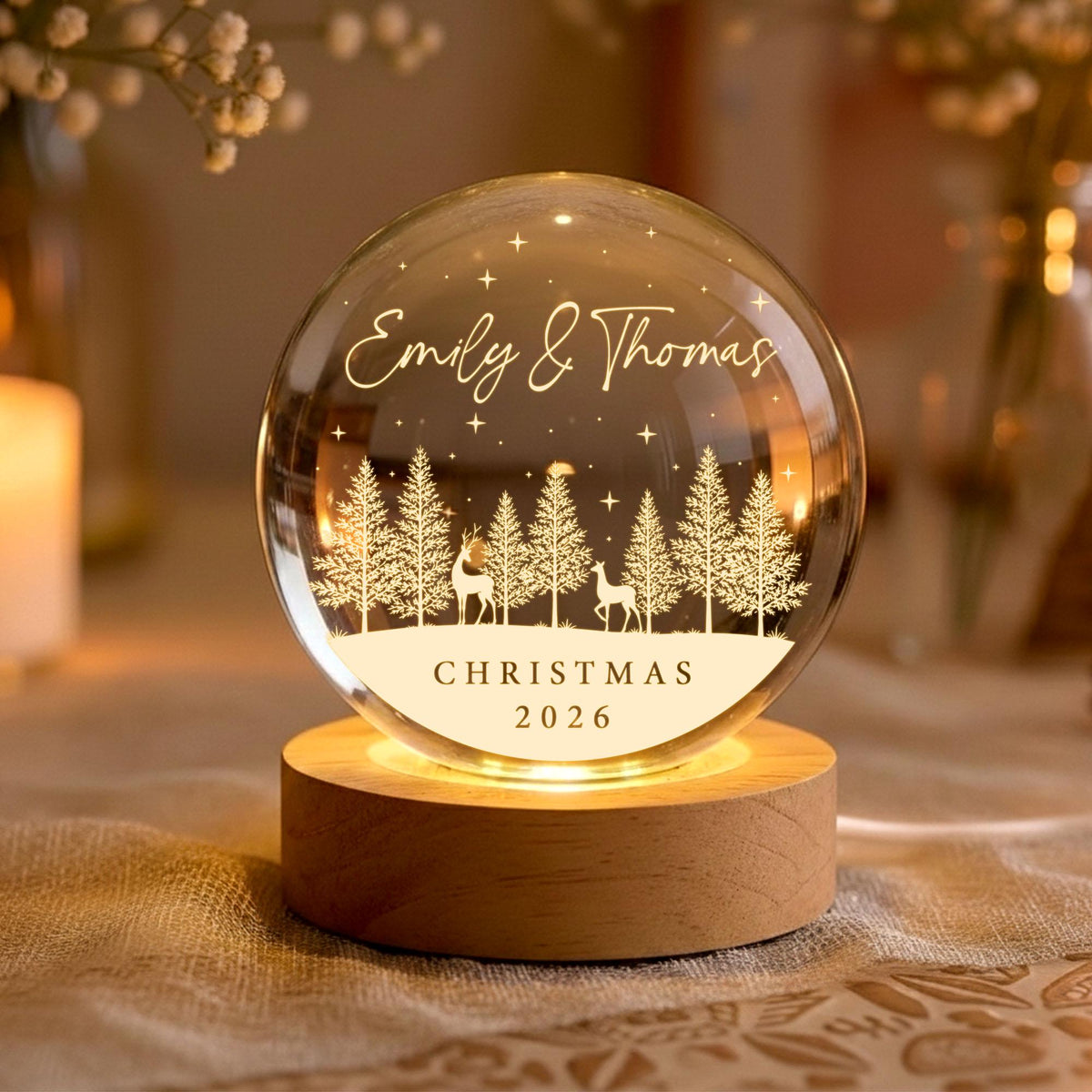 May include: A clear glass sphere night light on a round wooden base. The sphere features an etched design of a forest with two deer, stars, and the text "Emily & Thomas" at the top and "CHRISTMAS 2026" at the bottom, illuminated from below.