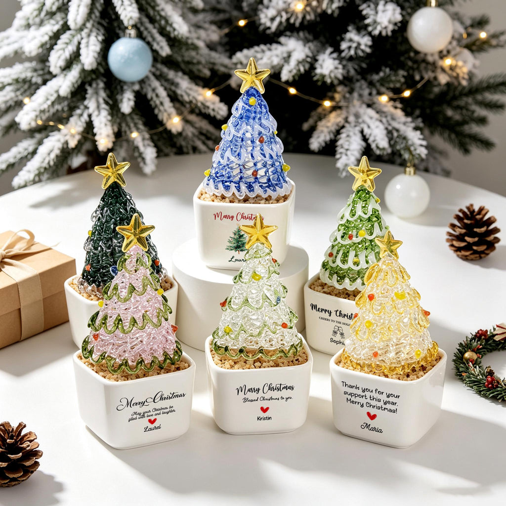 May include: Six miniature glass Christmas trees in white square ceramic pots. Each tree features a gold star topper and colorful glass accents. The pots are printed with Merry Christmas text and names like Laurel, Kristin, and Maria, along with small red heart icons.