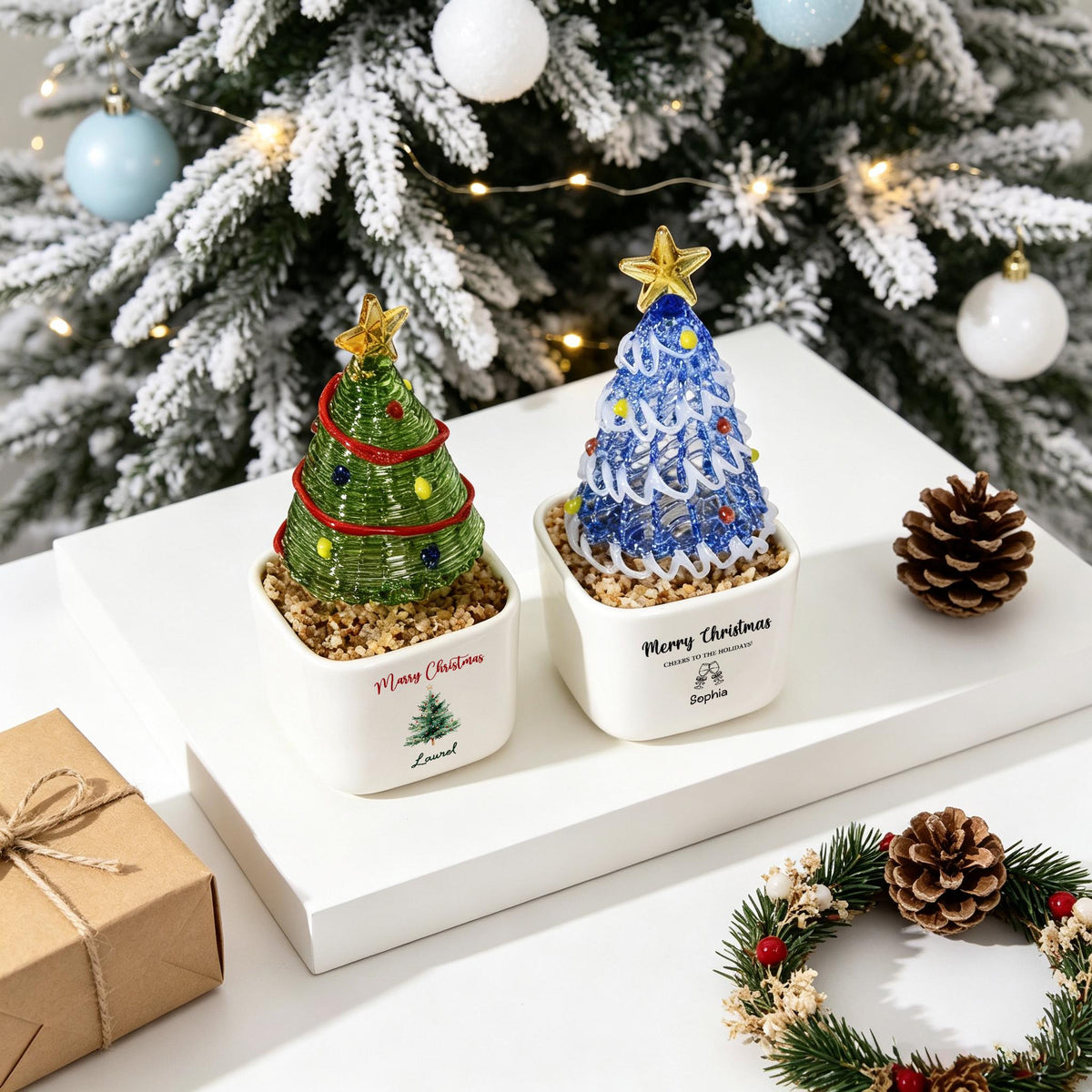 May include: Two glass Christmas tree ornaments in white square planters filled with small stones. One tree is green with a red garland and yellow lights, the other is blue with white garland and multi-colored lights. Both feature gold star toppers.