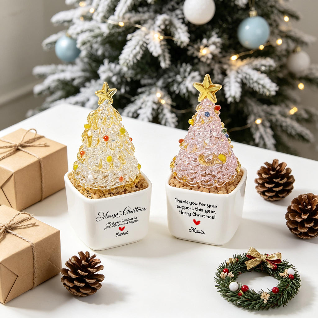 May include: Two decorative glass Christmas trees in white square ceramic pots. One tree is clear glass with colorful accents, the other is pink glass. The pots feature printed text including Merry Christmas, names Laurel and Maria, and red hearts.