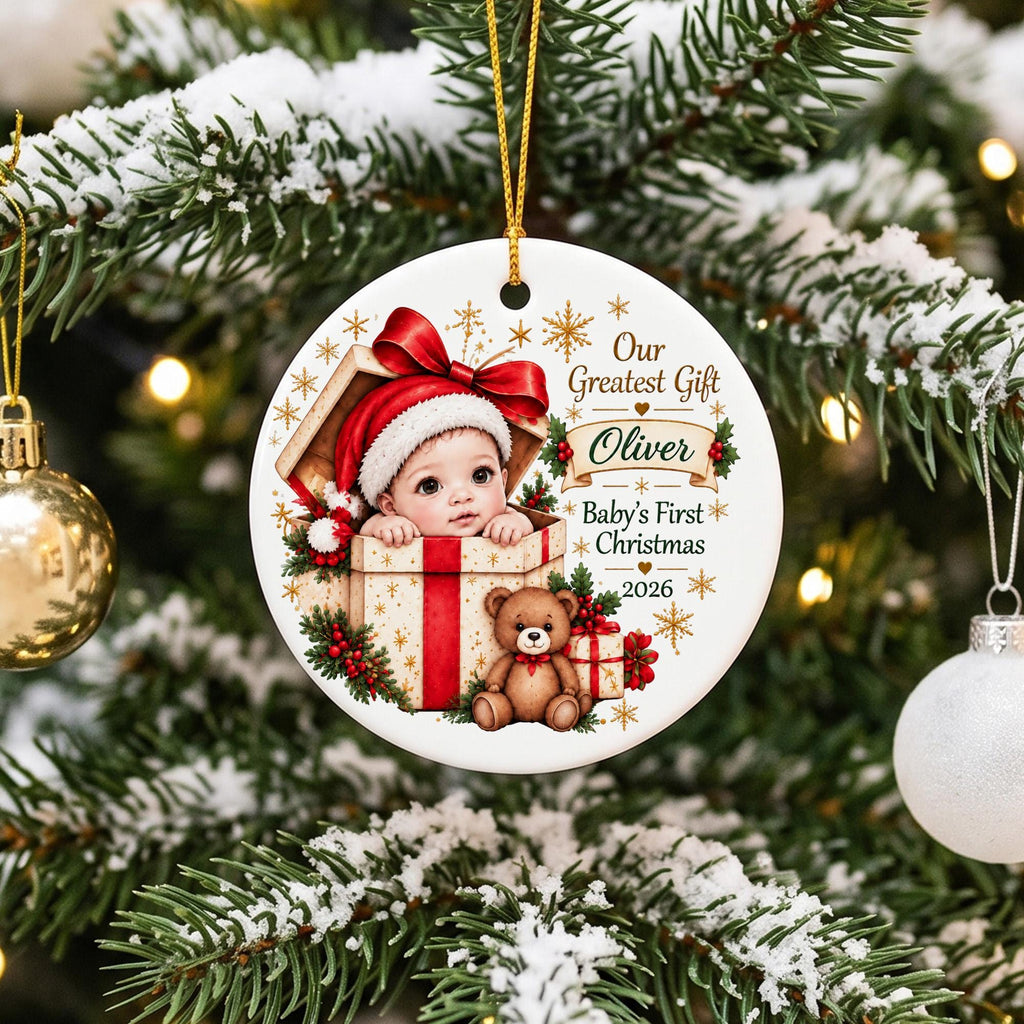 May include: A round white ceramic Christmas ornament featuring a baby in a Santa hat inside a gift box, a brown teddy bear, and the text Our Greatest Gift, Oliver, Baby's First Christmas, 2026, with gold star accents and a gold hanging cord.