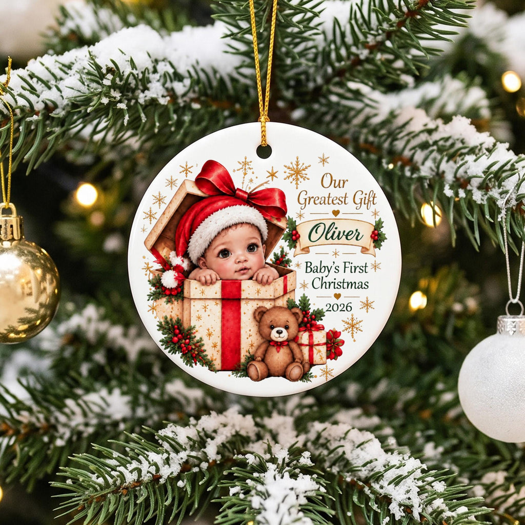 May include: A round white ceramic Christmas ornament featuring a graphic of a baby in a Santa hat inside a gift box with a teddy bear. The text reads Our Greatest Gift, Oliver, Baby's First Christmas, and 2026, accented with gold snowflakes.