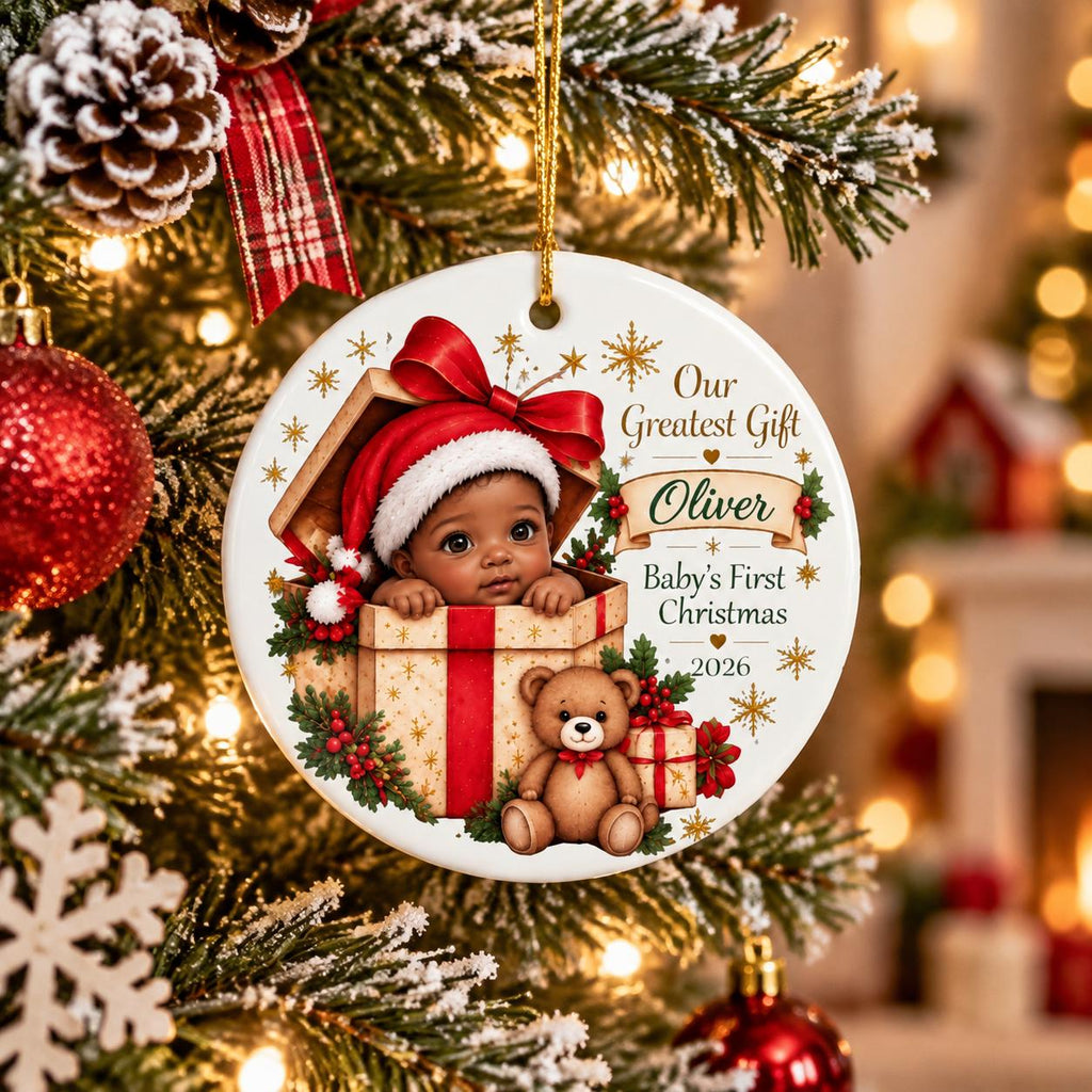 May include: A round white ceramic Christmas ornament featuring a baby in a Santa hat inside a gift box, a teddy bear, and the text Our Greatest Gift, Oliver, Baby's First Christmas, 2026. The design includes red ribbons, holly, and gold stars.