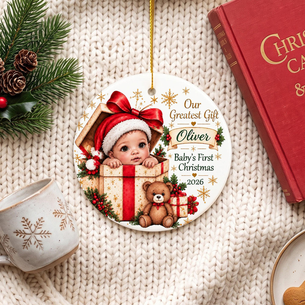 May include: A round white ceramic Christmas ornament featuring a baby wearing a red Santa hat inside a gift box, a brown teddy bear, and gold snowflake accents. The text reads: Our Greatest Gift, Oliver, Baby's First Christmas, 2026.