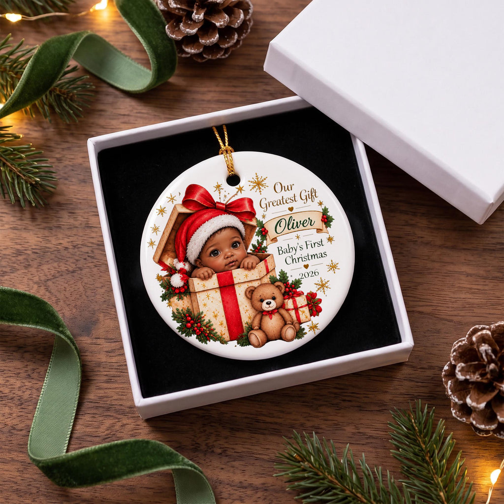 May include: A round white ceramic Christmas ornament featuring a graphic of a baby in a Santa hat inside a gift box with a teddy bear. The text reads Our Greatest Gift, Oliver, Baby's First Christmas, and 2026, surrounded by gold stars.